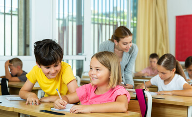 Fototapeta premium Kids sitting in classroom and performing test. Boy cheating and looking in classmate's exrcise book.