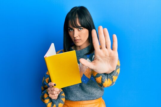 Young hispanic woman reading book with open hand doing stop sign with serious and confident expression, defense gesture