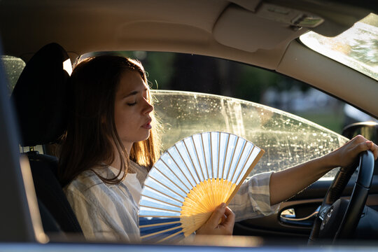 Exhausted young woman driver with hand fan suffering from heat in car, has problem with a non-working air conditioner, try to cool herself on hot sunny day. Summer season concept. 