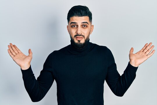 Handsome man with beard wearing turtleneck sweater clueless and confused with open arms, no idea and doubtful face.