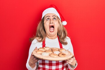 Middle age caucasian woman wearing baker apron and christmas hat holding tray with biscuits angry...