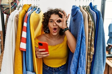 Young hispanic woman searching clothes on clothing rack using smartphone doing ok gesture shocked with surprised face, eye looking through fingers. unbelieving expression.