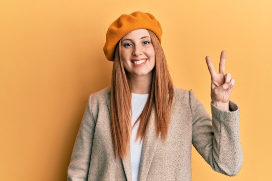 Young irish woman wearing french look with beret smiling with happy face winking at the camera doing victory sign. number two.