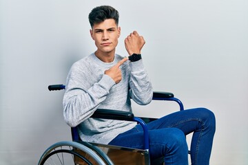 Young hispanic man sitting on wheelchair in hurry pointing to watch time, impatience, looking at...