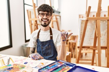 Hispanic man with beard at art studio smiling with happy face looking and pointing to the side with thumb up.