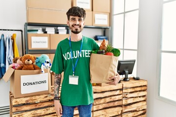 Hispanic man with beard wearing volunteer t shirt at donations point looking positive and happy...
