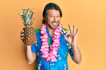 Middle age handsome man wearing hawaiian lei holding pineapple doing ok sign with fingers, smiling...