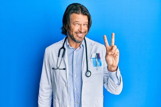 Middle age handsome man wearing doctor uniform and stethoscope smiling with happy face winking at the camera doing victory sign. number two.