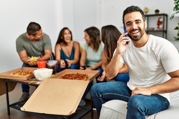 Group of young hispanic friends eating italian pizza sitting on the sofa. Man smiling happy and talking on the smartphone at home.