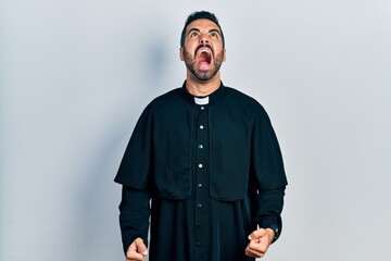 Handsome hispanic man with beard wearing catholic priest robe angry and mad screaming frustrated and furious, shouting with anger. rage and aggressive concept.