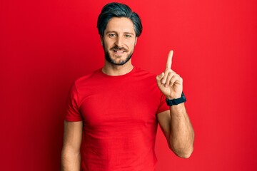 Young hispanic man wearing casual red t shirt showing and pointing up with finger number one while smiling confident and happy.