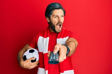 Young hispanic man football hooligan holding ball using tv control angry and mad screaming frustrated and furious, shouting with anger. rage and aggressive concept.