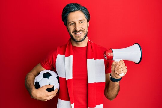 Young hispanic man football hooligan holding ball using megaphone smiling with a happy and cool smile on face. showing teeth.