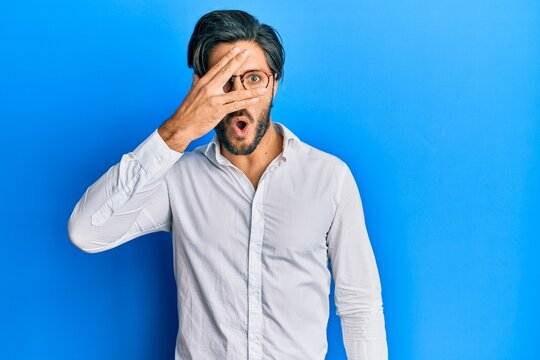 Young hispanic man wearing casual clothes and glasses peeking in shock covering face and eyes with hand, looking through fingers afraid