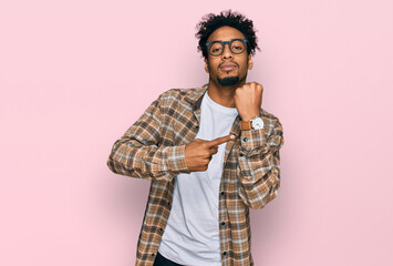 Young african american man with beard wearing casual clothes and glasses in hurry pointing to watch...