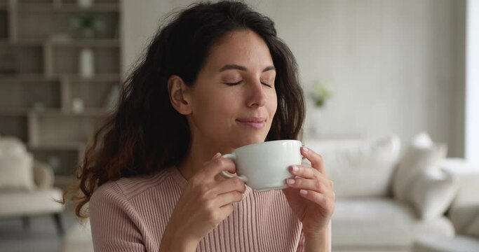 Happy pensive calm beautiful hispanic woman drinking cup of tea or coffee, daydreaming enjoying peaceful carefree morning time alone in living room, planning day or imagining future, resting indoors.