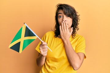 Young hispanic woman holding jamaica flag covering mouth with hand, shocked and afraid for mistake....