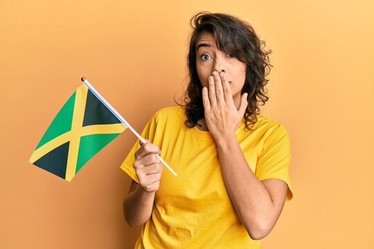 Young hispanic woman holding jamaica flag covering mouth with hand, shocked and afraid for mistake. surprised expression