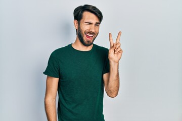Fototapeta premium Young hispanic man wearing casual white tshirt smiling with happy face winking at the camera doing victory sign. number two.