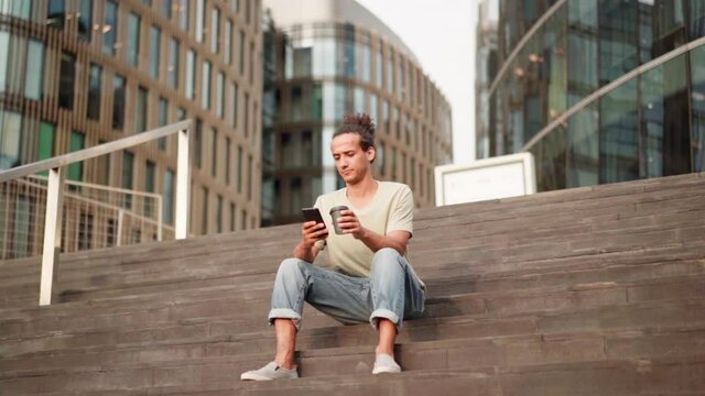 Zoom in slow motion shot of young man with coffee sitting on steps outdoors, using cell phone, browsing internet or text messaging, and thinking. Guy resting or waiting for smb