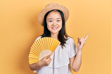 Young chinese girl waving hand fan cooling air in summer smiling happy pointing with hand and finger to the side