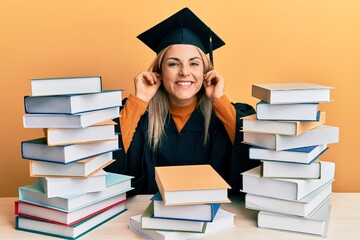 Young caucasian woman wearing graduation ceremony robe sitting on the table smiling pulling ears...