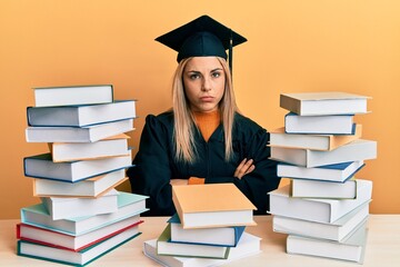 Young caucasian woman wearing graduation ceremony robe sitting on the table skeptic and nervous, disapproving expression on face with crossed arms. negative person.