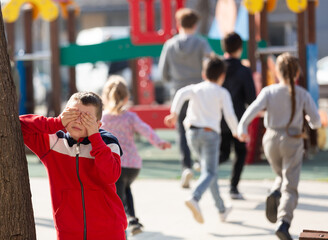 Portrait of preschool boy playing hide-and-seek with friends, covering his eyes with hands near big tree