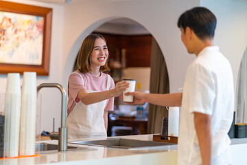 Young Asian woman coffee shop employee barista working at cafe. Smiling female waitress or cashier preparing takeaway order to customer. Small business owner and part time job working concept