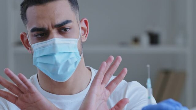 Woman doctor nurse holding syringe with vaccine disease remedy arabic hispanic man patient wears face mask says no vaccination immunization makes gesture with hands against injection denies disagrees