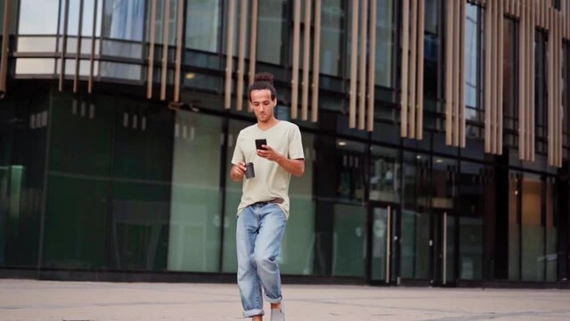 Panning slow motion shot of confident young man walking down city street, text messaging on cell phone, drinking takeaway coffee and smiling feeling carefree. Woman with laptop walking in background