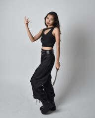Full length portrait of pretty young asian girl wearing black tank top, utilitarian  pants and leather boots. Standing pose holding a knife,  isolated against a  studio background.