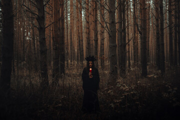 witch in a black costume holds candles in a dark forest