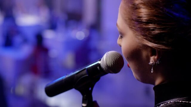 Rear or back view of a elegant woman singing to microphone on blur background . Glamorous singer holding a mike and singing blues . Beauty glamour fashion woman with microphone over restaurant hall