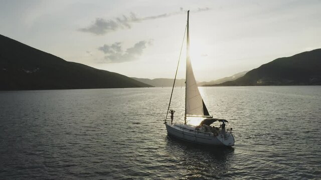 Evening romantic tracking shot of sailboat cruising on sea. Woman posing on bow deck. Water surface reflecting evening sun.