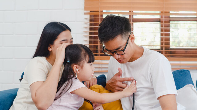 Happy Cheerful Asian Family Dad, Mom And Daughter Playing Funny Game As Doctor Having Fun On Sofa At Home. Self-isolation, Stay At Home, Social Distancing, Quarantine For Coronavirus Prevention.