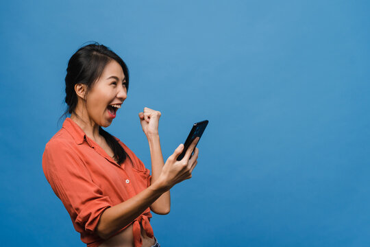 Surprised Young Asia Lady Using Mobile Phone With Positive Expression, Smiles Broadly, Dressed In Casual Clothing And Stand Isolated On Blue Background. Happy Adorable Glad Woman Rejoices Success.