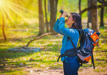 Woman wearing a blue jean and with backpack. she is using a binoculars in the forrest with sun rays.