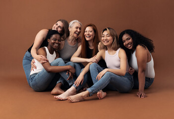 Portrait of six laughing women of different ages and body types sitting together on a brown...