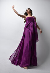 Full length  portrait of pretty brunette asian girl wearing purple flowing  gown. Standing pose in low angle on on studio background.