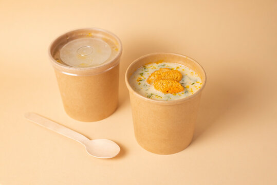 Creamy fish soup to go with salmon, croutons on light orange background. Healthy lunch. Take away food in brown paper craft container, wooden spoon. Concept eco restaurant delivery.