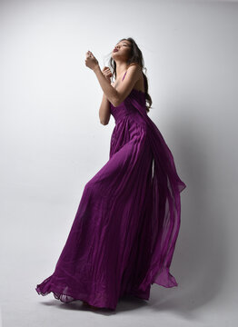 Full Length  Portrait Of Pretty Brunette Asian Girl Wearing Purple Flowing  Gown. Standing Pose In Low Angle On On Studio Background.