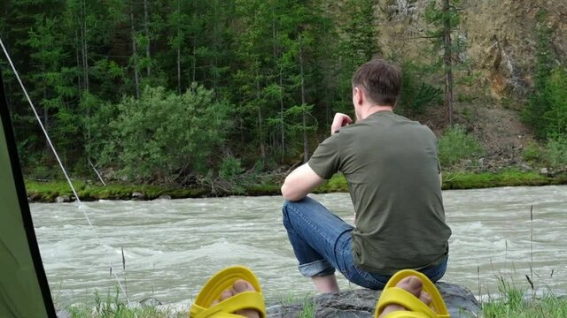 View from camping tent. A man sits on a stone on the bank of a mountain river. Slow motion