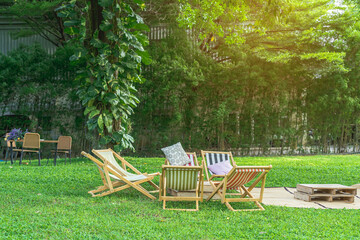 Many deck chairs and pillows with wooden table in the courtyard is surrounded by shady green grass. Comfortable pillows on outdoor patio chair and table in garden. Summer vacation. Selective focus.