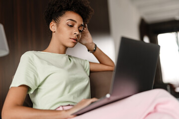 Young black woman working with laptop while sitting on armchair