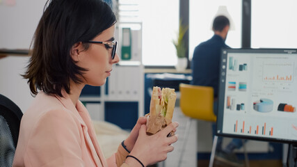 Entrepreneur woman eating tasty sandwich during takeout lunchbreak working at financial graphs in...