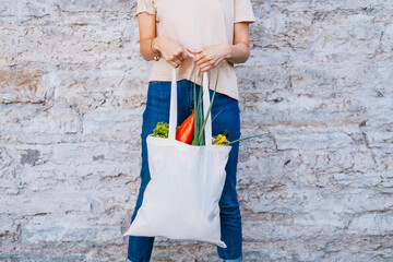 Woman holding white canvas tote bag with vegetables near brick wall.