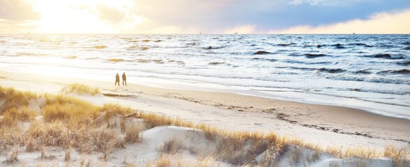 A couple is walking through the Baltic sea shore at sunset. Sand dunes and plants close-up. Colorful evening clouds. Waves and water splashes. Idyllic seascape. Latvia