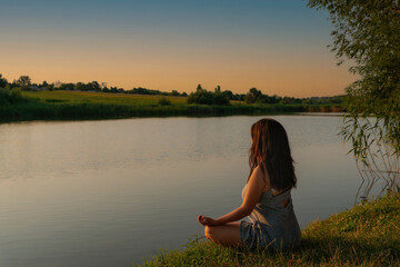 Woman meditating in lotus pose during sunset. Girl meditates on shore of river alone with herself. Yoga, meditation, tranquility, relax in nature. Beatiful landscape