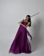 Full length  portrait of pretty brunette asian girl wearing purple flowing  gown. Sitting pose holding a sword  on on studio background.